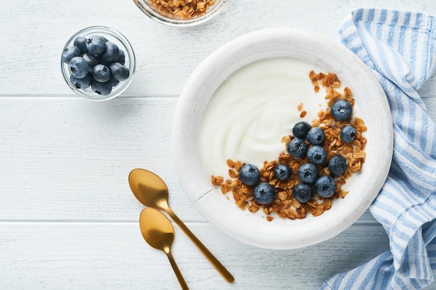 Yogurt Yogur griego con granola y arándanos frescos en un tazón blanco sobre fondo de madera blanca vieja Concepto de desayuno matutino Comida saludable para el desayuno vista superior