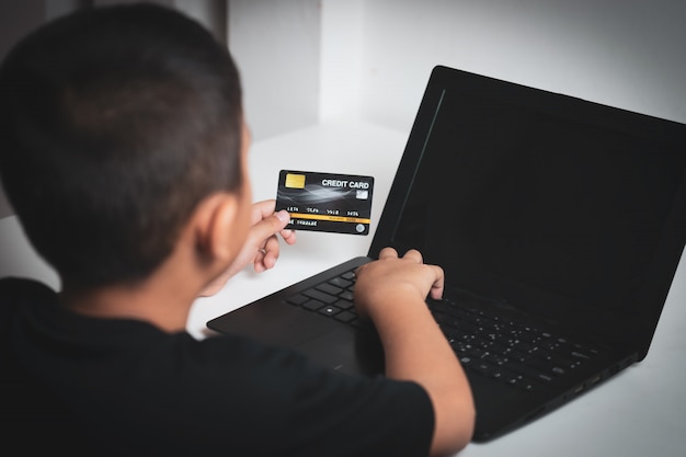 Foto yong asian children mit der gelben haut, schwarze kreditkarte, schwarzen laptop auf weißer tabelle halten.