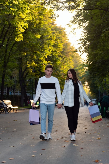 Yong pareja de enamorados está caminando en el parque cogidos de la mano con bolsas de la compra. Marco vertical.