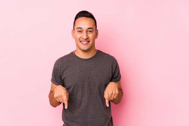 Yooung hombre latino posando en una pared rosa apunta hacia abajo con los dedos, sentimiento positivo.
