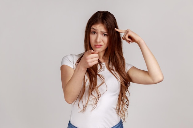 You39re idiota com plano louco insano Mulher descontente mostrando gesto estúpido e apontando para a câmera acusando de ideia idiota vestindo camiseta branca Foto de estúdio interior isolada em fundo cinza