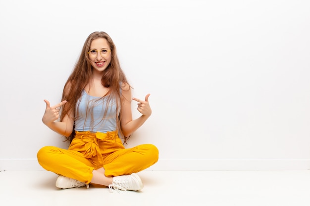 Yound mujer rubia que parece orgullosa, arrogante, feliz, sorprendida y satisfecha, apuntando a sí misma, sintiéndose como una ganadora sentada en el suelo