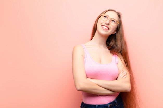 Yound mujer rubia sintiéndose feliz, orgullosa y esperanzada, preguntándose o pensando, mirando hacia arriba para copiar el espacio con los brazos cruzados