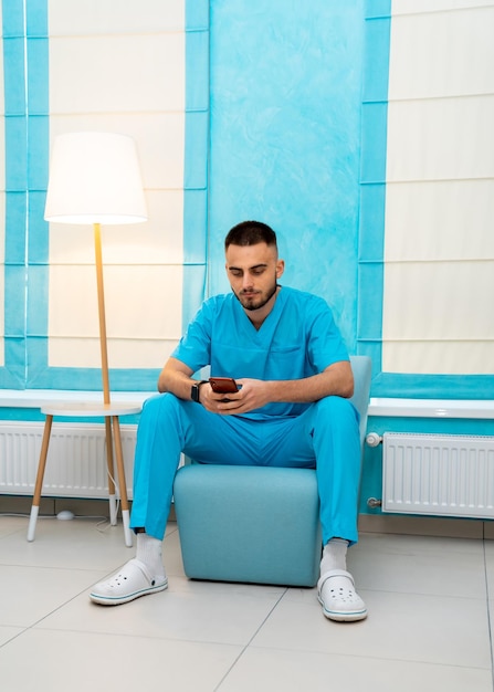 Younf lindo médico está navegando na internet no telefone após um dia pesado de trabalho. Médico se senta na cadeira com o telefone nas mãos.