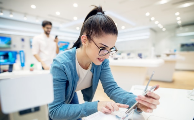 Young satisfizo a una chica hermosa, encantadora y elegante probando el nuevo modelo de un móvil desde el escritorio de la tienda de tecnología.