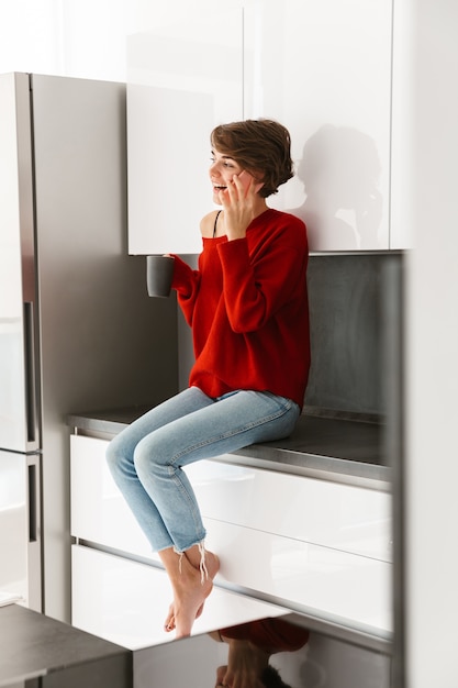 Younh sonriente mujer vistiendo suéter sentado en una mesa de la cocina en casa, mediante teléfono móvil