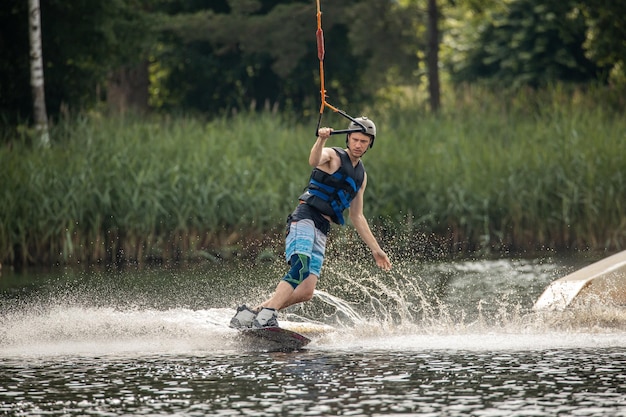 Ypung homem wakeboarding e fazendo diferentes truques de tabuleiro Wake boarding esportista homem saltando alto Wake boarding raley truque com enorme salpico de água no parque de cabo