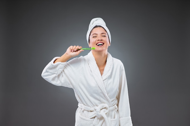Zähneputzen und Mundpflege. Ein lächelndes Mädchen in einem Bademantel und mit einem Handtuch, das nach dem Baden um den Kopf gewickelt ist und eine Zahnbürste in der Hand hält, die sich die Zähne putzt