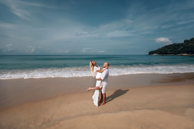 Zärtlich umarmt und zärtlich küssend genießt ein junges Paar in weißen Kleidern einen Hochsommertag an einem nassen Sandstrand. Phuket, Thailand.