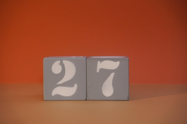 Zahl 27 auf grauen Holzwürfeln in Großaufnahme Konzept der Datums- und Uhrzeitangabe Mathe-Konzept Kopierraum für Text oder Ereignis Weiße Zahlen 27 Bausteine orangefarbener Hintergrund Selektiver Fokus