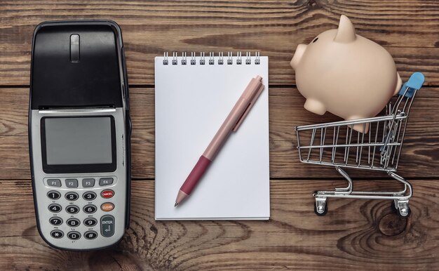 Zahlung POS-Terminal Einkaufswagen mit Sparschwein Notebook auf Holztisch Sparen Sie Geld Einkaufskonzept Ansicht von oben