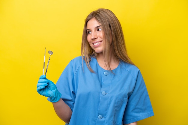 Zahnärztin, kaukasische Frau, die Werkzeuge isoliert auf gelbem Hintergrund hält, zur Seite schaut und lächelt