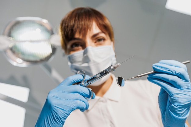 Zahnärztin mit blauer Gesichtsmaske und Handschuhen untersucht Zähne mit Spritze und Spiegelansicht vom Patienten