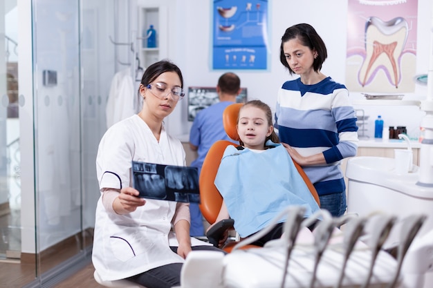 Zahnarzt, der die Röntgenaufnahme des kleinen Kindes untersucht, das auf einem Stuhl sitzt und ein zahnmedizinisches Lätzchen trägt. Stomatologe, der Mutter des Kindes in der Gesundheitsklinik mit Röntgenaufnahme die Zahndiagnose erklärt.