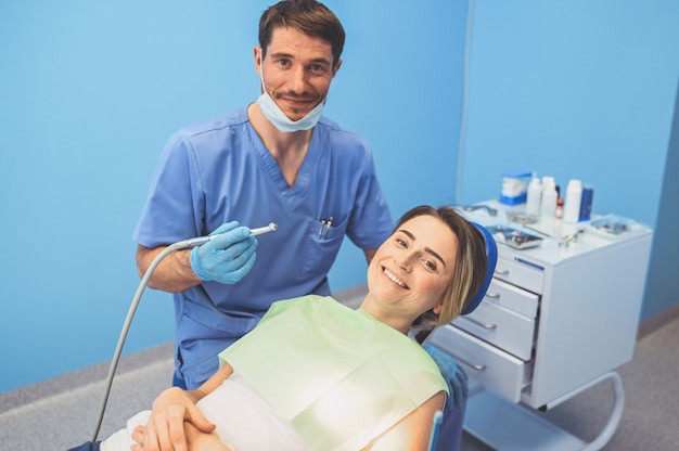 Zahnarzt, der die Zähne eines Patienten mit zahnärztlicher Ausrüstung in der Zahnarztpraxis untersucht. Stomatologie- und Gesundheitskonzept. Junger hübscher männlicher Arzt in der medizinischen Einweggesichtsmaske, lächelnde glückliche Frau