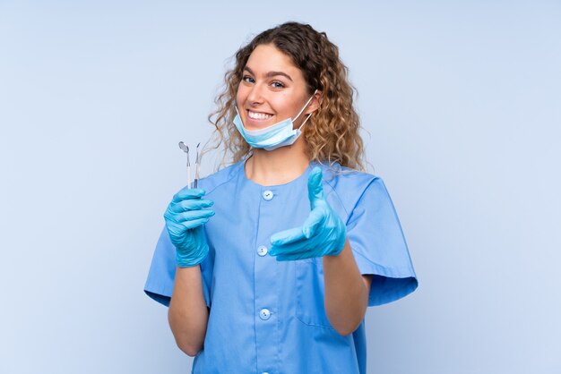 Zahnarzt der jungen blonden Frau, die Werkzeuge auf Handshaking der blauen Wand nach vielem Geschäft hält