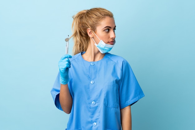 Zahnarzt der jungen Frau, der Werkzeuge über der blauen Wand hält, die Seite schaut