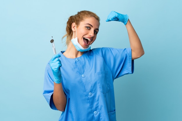 Zahnarzt der jungen Frau, der Werkzeuge über isoliertem Blau hält, macht starke Geste