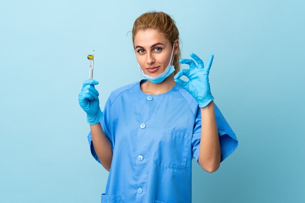 Zahnarzt der jungen Frau, die Werkzeuge über isolierter blauer Wand hält, die ein OK-Zeichen mit den Fingern zeigt