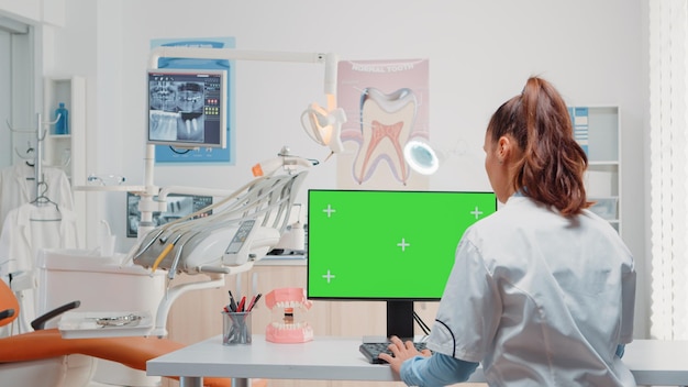 Zahnarzt mit Computer mit horizontalem grünem Bildschirm auf dem Display. Spezialist, der einen Monitor mit Chroma-Key und isolierter Vorlage für die Mundpflege betrachtet, während er mit zahnärztlichen Geräten assistiert