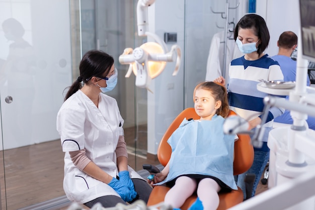 Zahnarzt mit Gesichtsmaske in der Kurskinderprüfung mit zahnärztlichem Lätzchen in der Zahnarztpraxis. Zahnmediziner während der Kinderhöhlenberatung in der Stomatologie-Büro mit moderner Technologie.