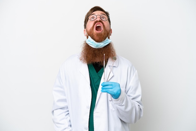 Zahnarzt rötlicher Mann mit Werkzeugen isoliert auf weißem Hintergrund nach oben schauend und mit überraschtem Ausdruck