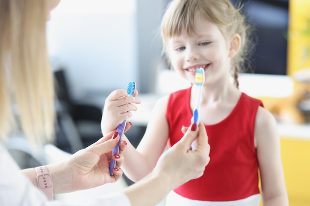 Zahnarztdoktor, der zwei Zahnbürsten vor der Nahaufnahme des kleinen Mädchens hält