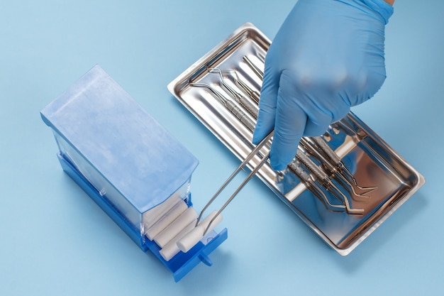 Zahnarzthand in einem Latexhandschuh mit Pinzette hält einen Baumwolltampon. Satz von zusammengesetzten Füllinstrumenten für die zahnmedizinische Behandlung in der Stahlschale auf dem Hintergrund. Medizinisches Werkzeugkonzept. Ansicht von oben.