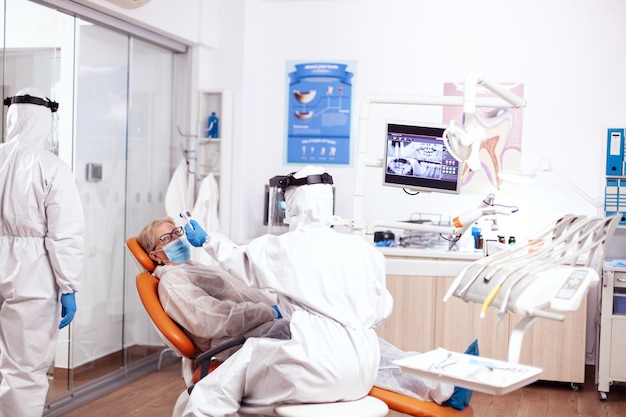 Zahnarztkrankenschwester, die die Temperatur des Patienten mit einem Gefahrstoffanzug mit einem digitalen Thermometer misst. Ältere Frau in Schutzuniform während der ärztlichen Untersuchung in der Zahnklinik.