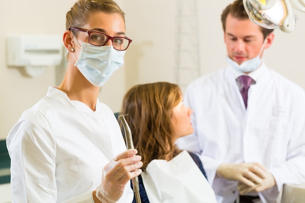 Zahnarztpraxis hält einen Bohrer und schaut auf den Betrachter, in dem ihre Kollegin einer Patientin eine Behandlung gibt