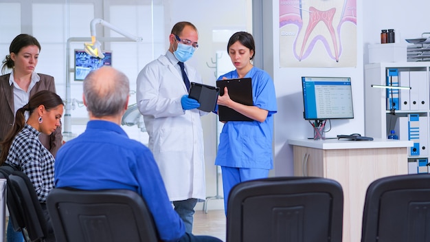 Zahnmediziner, der mit einem Assistenten im Wartezimmer der stomatologischen Klinik vor der Untersuchung der Patienten spricht, Krankenschwester, die Notizen in der Zwischenablage macht, während der Arzt auf dem Tablet schaut.