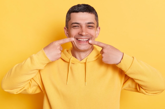 Zahnpflege Mundgesundheit Lächelnder gutaussehender brünetter Mann mit lässigem Hoodie, der auf seine perfekten Zähne zeigt und Zahn zeigt, nachdem er den Zahnarzt isoliert auf gelbem Hintergrund besucht hat