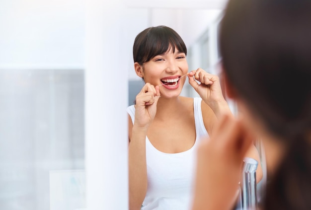 Zahnseide entfernt Aufnahme einer fröhlichen, attraktiven jungen Frau, die ihre Zähne mit Zahnseide behandelt, während sie zu Hause ihr Spiegelbild in einem Spiegel betrachtet