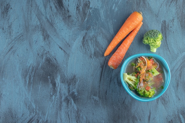 Zanahorias, brócoli y tazón de sopa de pollo, sobre fondo azul.