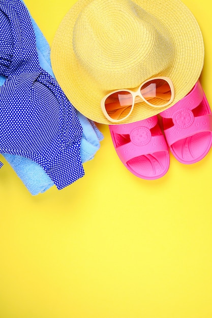 Zapatillas, bikini traje de baño, toalla, sombrero y gafas de sol sobre un fondo amarillo pastel.