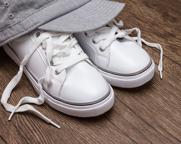 Zapatillas blancas con gorra gris sobre superficie de madera oscura. Estilo de moda deportiva