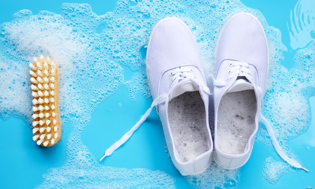 Zapatillas de deporte con espuma de detergente en polvo, disolución de agua y cepillo de madera sobre fondo azul. Lavado de zapatos sucios.