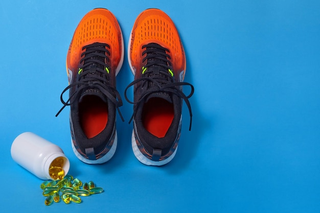 Zapatillas deportivas naranjas y pastillas de vitamina D y cápsulas de omega 3 derramadas de un frasco sobre un fondo azul