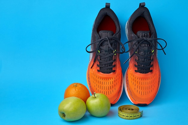 Foto zapatillas naranjas cinta métrica y frutas manzanas y naranjas sobre un fondo azul.