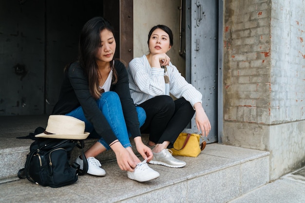 Zapato de corbata de turista asiática sentada cerca de una amiga en una antigua fábrica de monumentos con felicidad en verano mientras viaja. grupo de jóvenes japonesas se relajan en el suelo con mochila al lado de turismo.