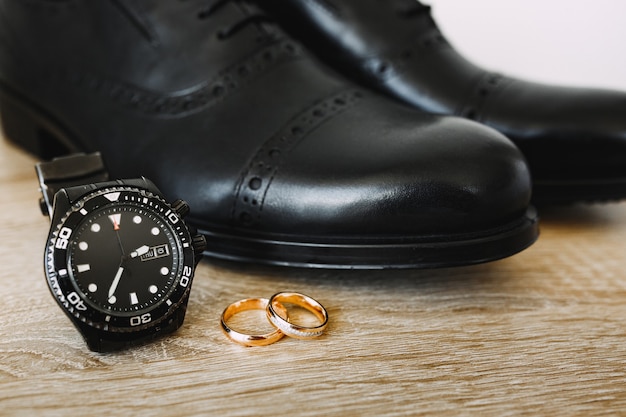 Zapatos de hombre negros en el suelo con anillos de boda de oro y relojes de pulsera mecánicos