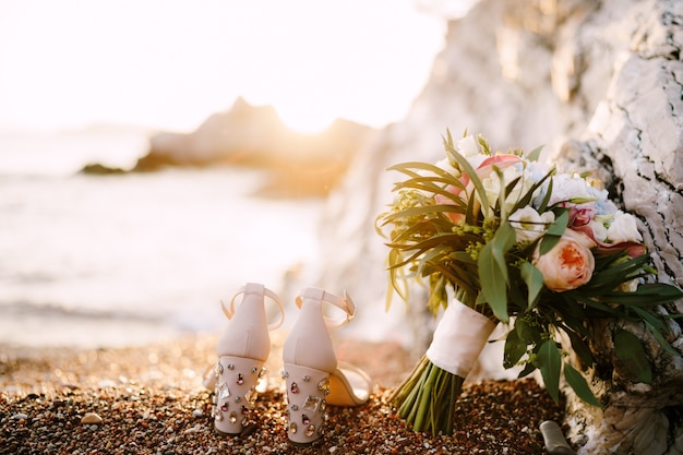 Zapatos de novia con pedrería en tacones de pie en la playa de guijarros de mar junto a un ramo de novia