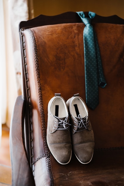 Zapatos de novio y corbata de lunares en una silla de cuero en la habitación.