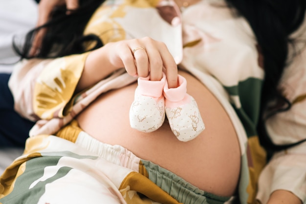 Zapatos pequeños para el bebé nonato en el vientre de la mujer embarazada. Mujer embarazada que sostiene los pequeños zapatos de bebé que se relajan en casa en dormitorio. Zapatos pequeños para el bebé nonato en el vientre de la mujer embarazada.