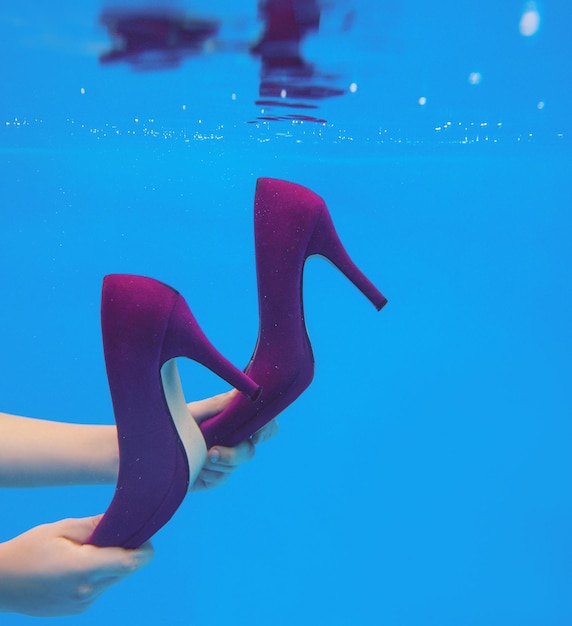 Zapatos de terciopelo violeta en manos de mujer bajo el agua en la piscina sobre fondo azul.