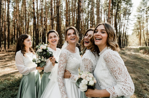 Zarte Braut mit lächelnden Brautjungfern in langen eleganten Kleidern, Brautjungfern mit glücklicher Braut am Hochzeitstag. Hochzeitssträuße werden von Freundinnen gehalten, Hochzeitstag,