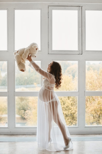 Zarte schwangere Frau im weißen Kleid, das mit Teddybär aufwirft