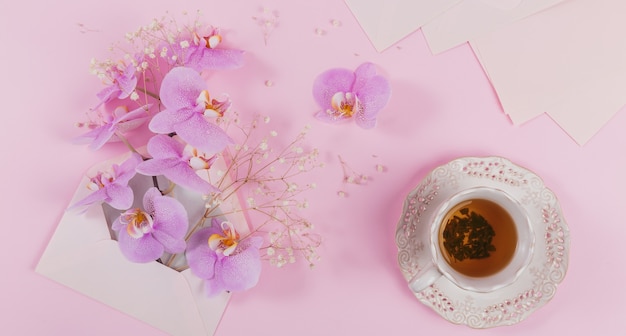 Zarte überkopfkomposition mit morgendlicher tasse tee, rosa brieftasche voller lila orchideenblüten und leerem umschlag auf hellrosa oberfläche