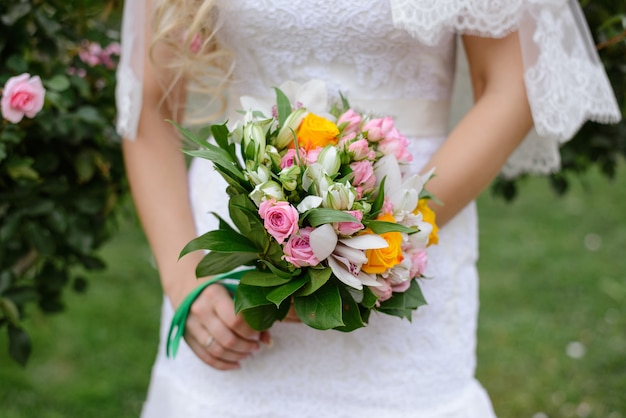 Zarter schöner Hochzeitsstrauß aus rosa und gelben Rosen und Orchideen mit viel Grün in den Händen der Braut hautnah