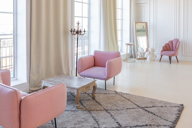 Zartes und gemütliches helles Interieur des Wohnzimmers mit modernen stilvollen Möbeln in Pastellrosa und weißen Wänden mit Stuckleisten bei Tageslicht